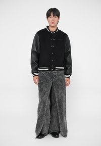Giacca varsity nera con maniche in pelle, polsini a strisce bianche, abbinata a pantaloni in denim grigi lunghi e strappati. Il modello si trova in piedi contro uno sfondo neutro.