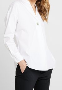 Weiße Baumwollbluse mit V-Ausschnitt, langen Ärmeln, Knopfdetails vorne und lockerer Passform, kombiniert mit schwarzen Hosen.