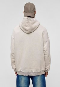 Beige oversized hoodie met textuur in de capuchon, geribbelde zoom en cuffs. Draagt het met vervaagde blauwe jeans en een lichtgrijze pet.