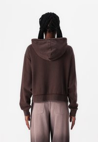Felpa corta marrone in materiale morbido con cappuccio regolabile, caratterizzata da maniche lunghe e un fit rilassato. Indossata con pantaloni grigio chiaro sbiaditi.