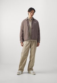 Light brown puffer jacket with a collar and logo, layered over a beige shirt. Loose beige trousers and white sneakers complete the outfit.