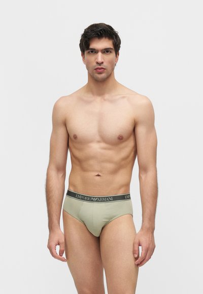 Μυώδης άνδρας στέκεται μπροστά σε απλό φόντο φορώντας ανοιχτό πράσινα εσώρουχα Emporio Armani με μαύρη ζώνη με λογότυπο.