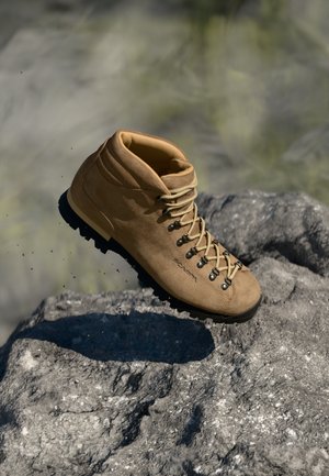 Beige semsket fjellstøvler med snøring foran, forsterket ankel og strukturelt gummisåle. Har metalløye og merkevare på siden.