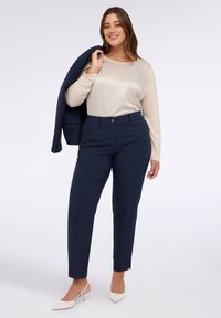 Marineblaue Polka-Dot-Hose mit taillierter Passform, kombiniert mit einem beigefarbenen Langarmoberteil. Das Modell hält einen passenden Blazer. Weiße spitze Schuhe.