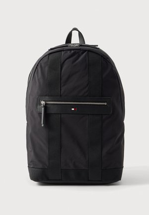 Sac à dos en tissu noir avec deux sangles verticales à l’avant, une poche avant zippée, une poignée supérieure et un petit logo rouge, blanc et bleu.