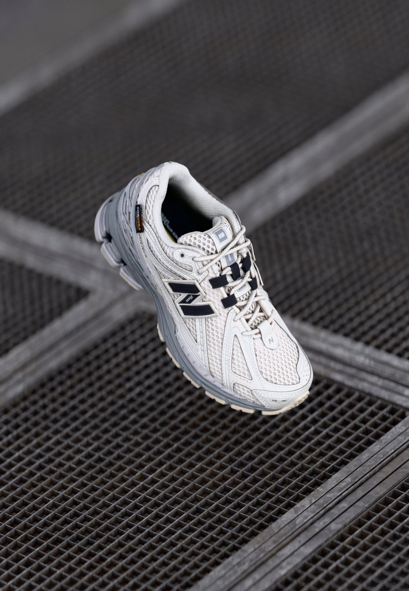 Hvide sportssko med mesh-overdel, grå detaljer, sorte snørebånd, polstret sål og struktureret gummisål. Brandlogo vist på siden.