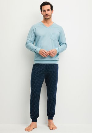 Un homme est debout pieds nus contre un mur blanc, portant une chemise bleu clair à manches longues et un pantalon de pyjama bleu marine à pois, les mains lâchement jointes.