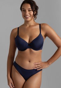 Χαμογελαστή γυναίκα με σγουρά σκούρα μαλλιά φοράει μπλε σουτιέν και ταιριαστό εσώρουχο, με φόντο μονόχρωμο γκρι.