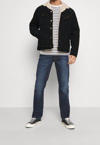 Sort denimjakke med beige fleecekrave, stribet hvid og marineblå skjorte, blå jeans og sorte sneakers med hvide såler.