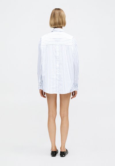 Άτομο με κοντά ξανθά μαλλιά, που φοράει μακρύ, λευκό πουκάμισο με κάθετες ρίγες και μαύρα παπούτσια, στέκεται με την πλάτη προς τον θεατή σε απλό φόντο.