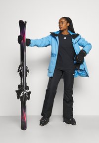 Blå skijakke med svarte detaljer, svart skjorte, svarte skibukser og svarte skistøvler. Holder lilla og svarte ski med festene.