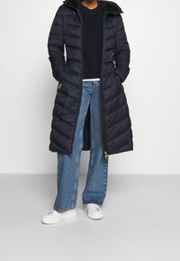 Langer, marineblauer Steppmantel mit Reißverschluss vorne, Kapuze und diamantförmiger Steppoptik; kombiniert mit ausgestellten Jeans und weißen Sneakern.