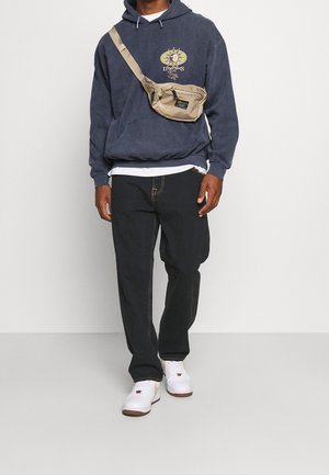 Felpa navy con design grafico, sovrapposta a una t-shirt bianca. Borsa a tracolla beige, pantaloni neri e scarpe bianche con dettagli dorati.