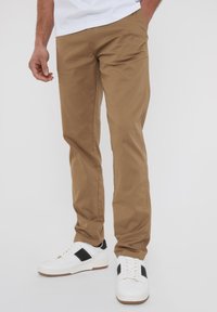 Krémové elastické kalhoty s užším střihem, mají hladkou texturu a rovný střih nohavic, doplněné bílými teniskami s černými akcenty.