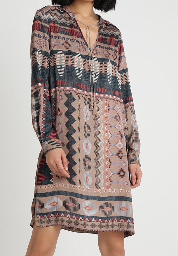 Femme portant une robe à manches longues, longueur genoux, avec des motifs géométriques et d'inspiration tribale dans des tons sourds de brun, noir, rouge et beige.
