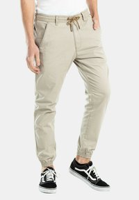 Pantalones jogger beige con cintura elástica, cordón, bolsillos laterales y puños ajustados. Combinados con zapatillas negras con detalles blancos.