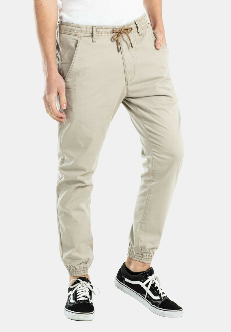 Pantalones jogger beige con cintura elástica, cordón, bolsillos laterales y puños ajustados. Combinados con zapatillas negras con detalles blancos.