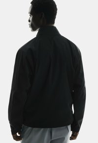 Black jacket with a high collar, smooth texture, and relaxed fit. Features long sleeves and minimalistic design, shot from a rear angle.