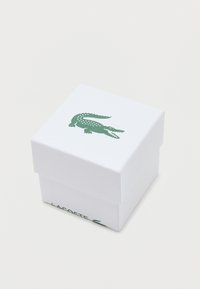 Bílá obdélníková krabička s texturovaným povrchem, na vrchu s zeleným krokodýlím logem a s nápisem "LACOSTE" vytištěným na jedné straně.