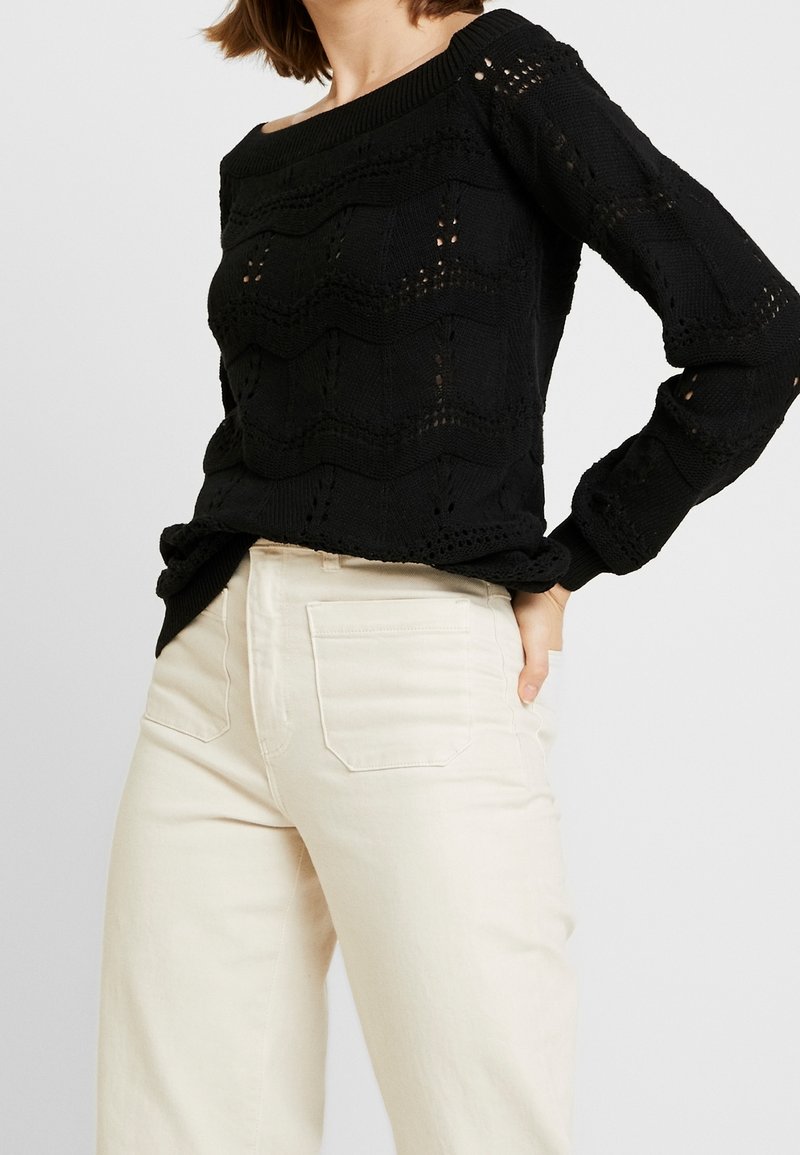 Femme portant un pull à manches longues noir en maille avec un motif ondulé et un pantalon taille haute blanc cassé, une main posée sur la hanche.
