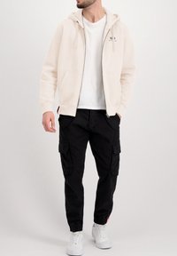 Lichtbeige hoodie met rits en voorzakken, gedragen over een wit T-shirt, gecombineerd met zwarte cargobroek en witte sneakers.