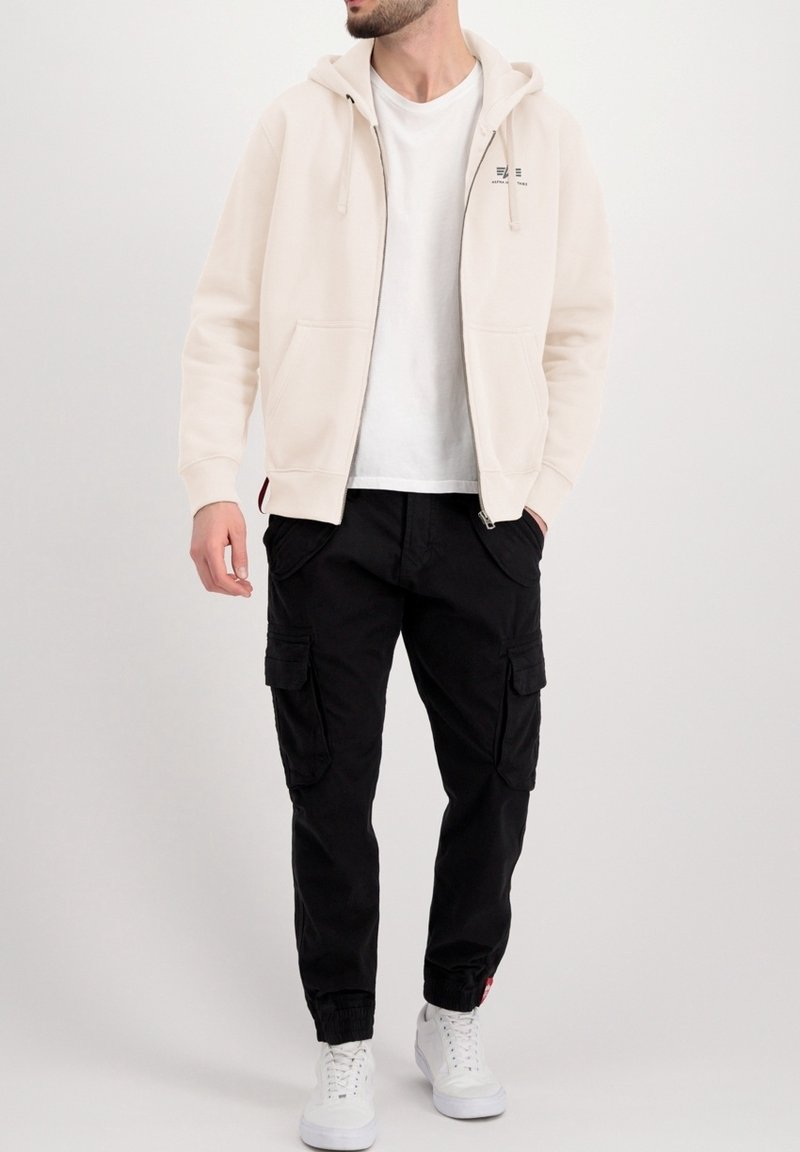 Lichtbeige hoodie met rits en voorzakken, gedragen over een wit T-shirt, gecombineerd met zwarte cargobroek en witte sneakers.