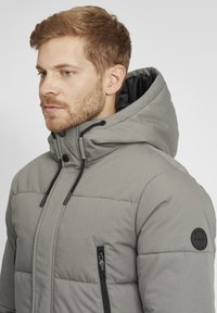 Veste isolée grise avec capuche, dotée d'une fermeture éclair à l'avant, de deux poches zippées et d'un badge sur la manche gauche. Texture lisse et design matelassé.