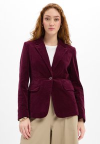 Jonge vrouw met lang rood haar draagt een bordeauxrode fluwelen blazer over een wit shirt en beige broek tegen een witte achtergrond.