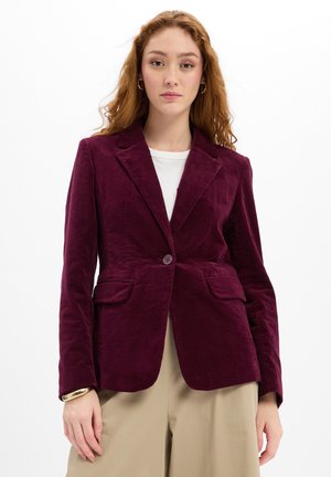 Young woman with long red hair wears a burgundy velvet blazer over a white shirt and beige pants against a white background.