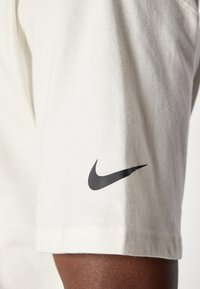 Chemise à manches courtes de couleur crème, en coton, avec un logo Nike noir proéminent sur la manche gauche. Texture lisse et design décontracté.