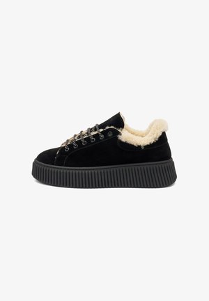 Zapatilla de ante negra con un forro de crema esponjoso, suela plana negra y ojales en tono plateado. Presenta una suela texturizada acanalada y cordones redondos.