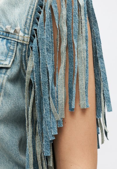 Υφάσμα τζιν με ξεφτισμένες λωρίδες που κρέμονται σαν φούντα δίπλα σε ένα χέρι, με μεταλλικά τρουκς κοντά στη ραφή της τσέπης.