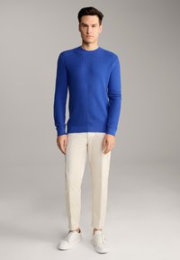 Pull en maille texturée bleu avec col rond, associé à un pantalon slim clair et des baskets blanches. Design simple et moderne.