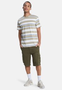 Jonge man staat met zijn handen in de zakken, gekleed in een beige gestreepte T-shirt, olijfgroene cargo shorts, witte sokken en beige sneakers.
