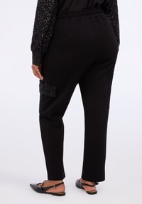 Pantaloni neri con una texture liscia, vita elastica e una patch decorativa in paillettes nere sul lato sinistro. Profilo elegante e a taglio dritto.