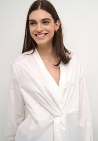 Camicia avvolgente bianca in cotone liscio, con scollo a V profondo, maniche lunghe e dettaglio annodato in vita.