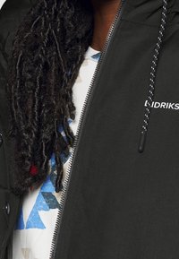 Zwarte jas met rits en een gladde textuur, voorzien van een capuchon en trekkoord. Wit logo op de borst. Daaronder een blauw-wit patroonhemd.