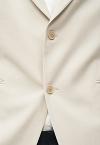 Crèmekleurige blazer met twee beige knopen vastgemaakt over een wit overhemd en donkere broek, met revers en zijzakken zichtbaar.