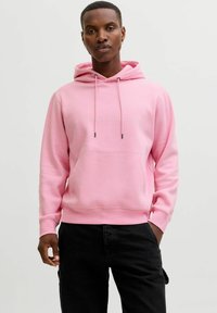Hoodie rose avec une poche kangourou, un cordon de serrage ajustable, et un texte de marque en blanc. Le tissu a une texture lisse et douce. Porté avec un pantalon noir.