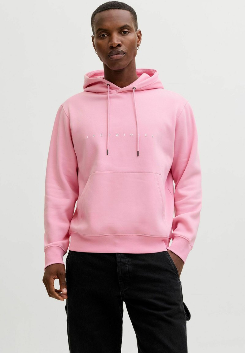 Hoodie rose avec une poche kangourou, un cordon de serrage ajustable, et un texte de marque en blanc. Le tissu a une texture lisse et douce. Porté avec un pantalon noir.