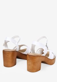 Sandali in faux leather bianchi con piattaforma, dotati di fasce larghe e fibbia alla caviglia regolabile. Tacco in legno naturale con texture simile al legno, alto circa 7 cm.
