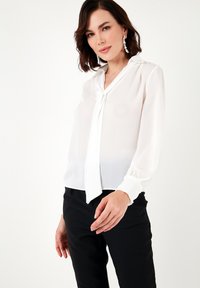 White blouse with a v-neck and tie detail. Made of lightweight, sheer fabric. Long sleeves with gathered cuffs. Paired with black tailored pants.