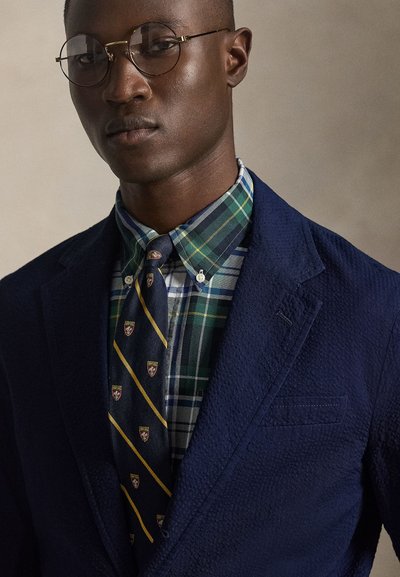 Άνδρας που φοράει στρογγυλά γυαλιά, μπλε σακάκι με υφή, πράσινο πουκάμισο με καρό μοτίβο και μπλε γραβάτα με μικρό έμβλημα και κίτρινες διαγώνιες ρίγες.