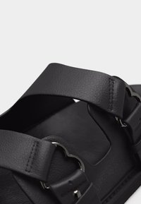 Μαύρο δερμάτινο σανδάλι με ρυθμιζόμενους ιμάντες και διακοσμητική ασημένια πόρπη σε σχήμα καρδιάς.