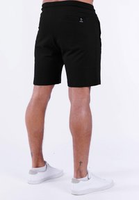 Schwarze Shorts mit elastischem Bund, versehen mit einer Gesäßtasche und einem Logo-Tag. Tragen kombiniert mit weißen Sneakers auf hellem Hintergrund.