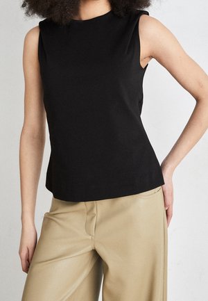 Vrouw met een mouwloos zwart topje en beige wijde broek, staand met één hand op haar heup tegen een effen achtergrond.