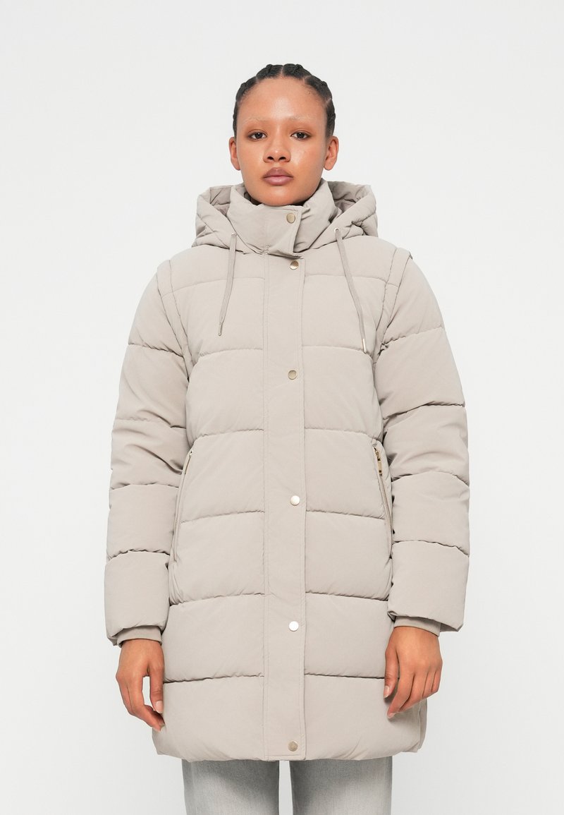 Parka matelassée gris clair avec capuche, dotée de poches latérales zippées et de boutons-pression, texture douce et design matelassé.