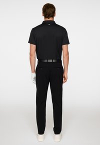 Svart pikétröja med korta ärmar, med krage och logotyp bak. Tillsammans med svarta byxor och vita sneakers. Golfhandske synlig.