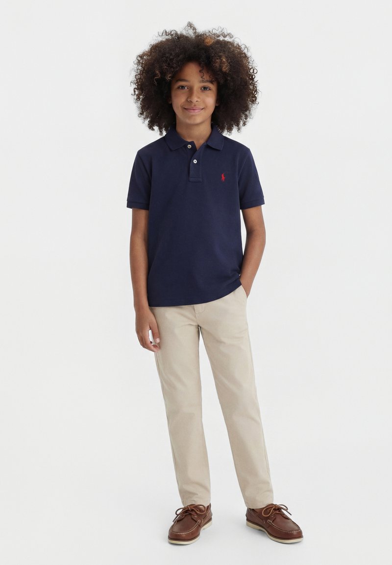 Fiú göndör hajjal, tengerészkék pólóingben, bézs nadrágban és barna fűzős cipőben, egy kézzel a zsebében áll.