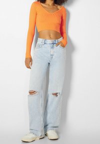 Pomarańczowy prążkowany crop top z długim rękawem w zestawie z jasnoniebieskimi spodniami dżinsowymi o wysokim stanie i szerokich nogawkach, z wystrzępionymi kolanami oraz białymi trampkami.